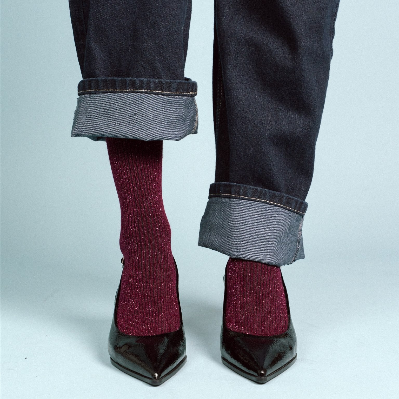 Person wearing dark jeans with rolled-up cuffs and burgundy socks with black shoes on a light gray background